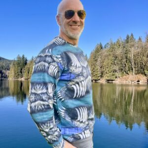 Male model stands in front of a blue lake and evergreen trees. Model is wearing Bear Claw Print Sweatshirt, and holds a blue waterbottle, as he stands striking and bold in texture and pattern. Adding depth to the model. Greens, Black, white, and blue palette of West Coast BC.