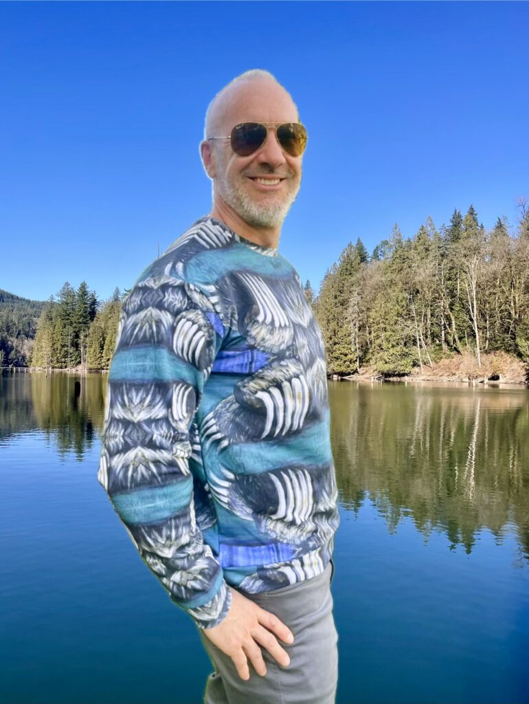 Model in the Bear Claw Sweatshirt by Tanya Tee Art standing by a West Coast lake and evergreens, showcasing the hand‑painted bear claw design.