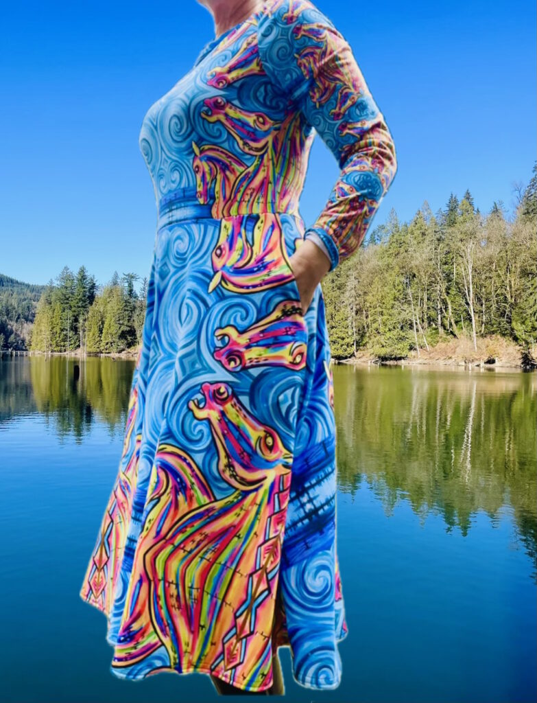 Model wears Killa Kelpies Dress showcasing side angle with hand in pocket. Swirling blues and bold colours construct Horse heads. Inspired by the Kelpies, Scotland.